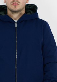 Giacca a cappuccio blu navy con zip frontale, texture soffice e foderatura verde a contrasto visibile lungo l'interno del cappuccio.