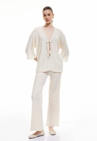 Haut en maille texturée blanche avec des manches larges et un lien à l'avant, associé à un pantalon large assorti. L'ensemble a une coupe décontractée et un tissu doux.