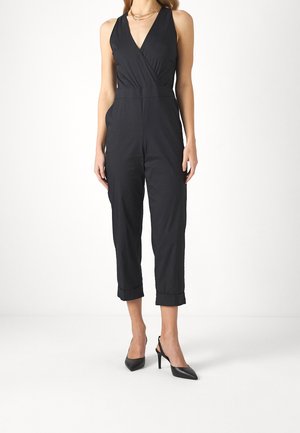 Kvinde iført sort ærmeløs jumpsuit med V-hals og bukser med ombuk ved anklerne, kombineret med sorte spidse høje hæle og en guldkæde.