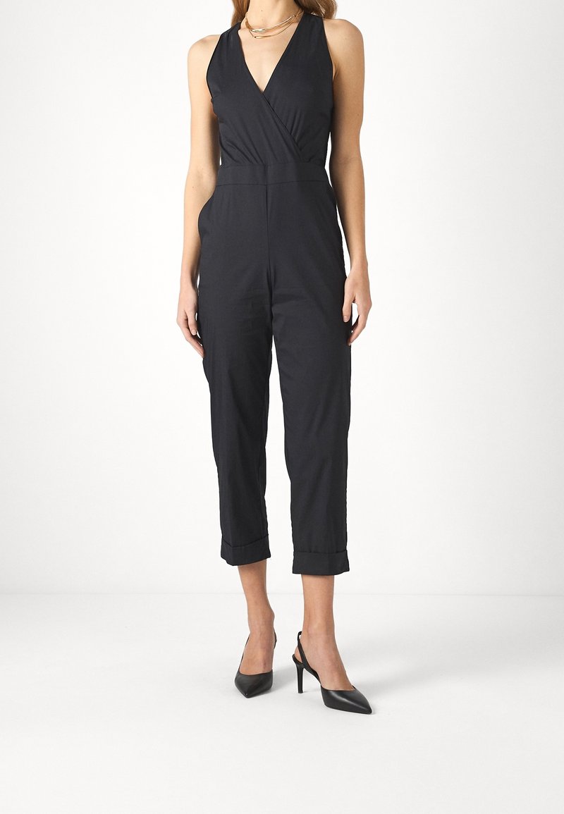 Femme portant une combinaison noire sans manches avec un col en V et un pantalon à revers aux chevilles, associée à des escarpins noirs pointus et un collier en or.