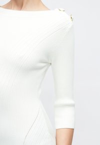 Λευκό τοπ από ριμπ πλεκτό με μανίκια τριών τετάρτων και χρυσαφένια κουμπιά στον ώμο, φορεμένο από ένα άτομο σε απλό φόντο.