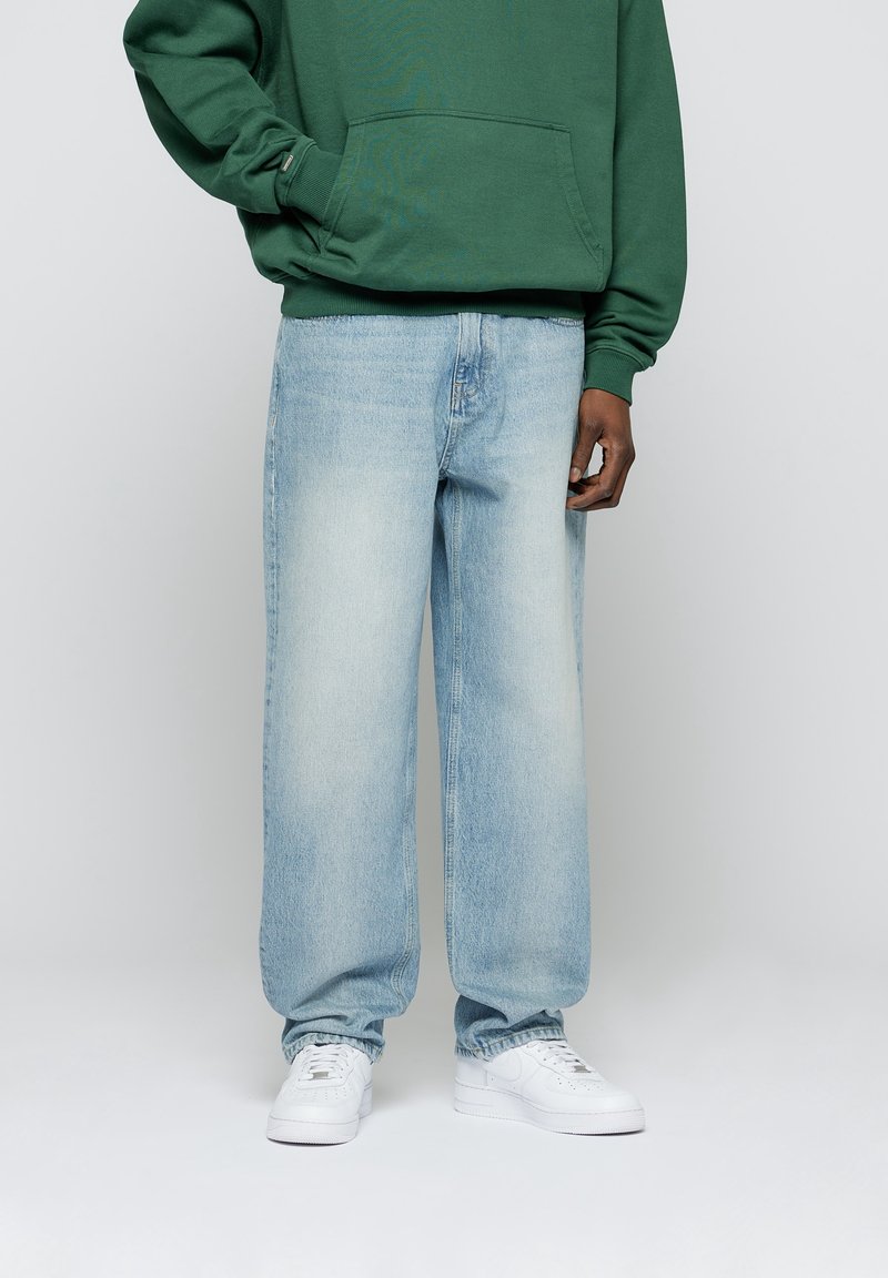 Personne portant un jean large bleu clair, des baskets blanches et un sweat-shirt vert les mains dans les poches, debout devant un fond uni.