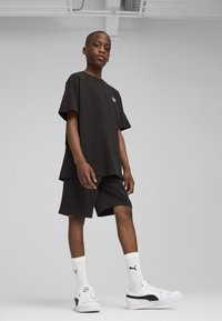 Ensemble en coton noir composé d'un t-shirt et d'un short, avec une coupe ample. Porté avec des chaussettes blanches de type crew arborant un logo noir et des baskets blanches avec des accents noirs.