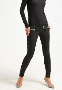 Pantalones negros ajustados con bolsillos con cremallera plateada, hechos de una tela suave; combinados con una blusa negra de manga larga y tacones metálicos.