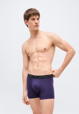 Jeune homme aux cheveux bruns courts portant un boxer Puma violet foncé, debout devant un fond gris clair, regardant vers la gauche.