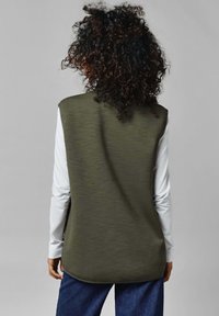 Olijfgroene mouwloze top met een textuurafwerking, gedragen over een wit shirt met lange mouwen, gecombineerd met blauwe jeans. Rond gesneden achterzoom.