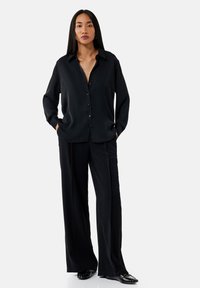 Camicia nera a maniche lunghe con design button-up e colletto, abbinata a pantaloni neri a gamba larga. Scarpe nere lucide completano il look.
