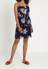 Robe bleu marine à motif floral avec encolure et taille à volants, dotée de bretelles spaghetti. Associée à des sandales plates rouges.