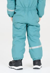 Teal barnoverall med elastiska ärmslut, utrustad med en reflexrand på varje ben, tillsammans med svarta vinterstövlar med vita sulor.