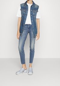 Chaleco de mezclilla sin mangas con botones en la parte delantera, combinado con una camiseta blanca, jeans ajustados de color azul claro y zuecos azul claro.