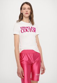 Bílý tričko s výrazným růžovým nápisem "VERSACE JEANS COUTURE", kombinované s lesklými růžovými šortkami se značkováním na stehně a texturovaným pasem.