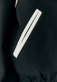 Czarna kurtka z tkaniny, z ukośnym białym paskiem na kieszeni, o gładkiej fakturze i marszczonym wykończeniu na dole.