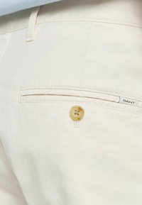 Crèmekleurige broeken met een gladde textuur, voorzien van een horizontale achterzak en een ronde, lichtbruine knop met bloemmotief.