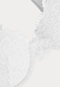 Soutien-gorge en dentelle blanche avec des motifs floraux, des bords scallopés détaillés, des bonnets lisses et une bande douce. La texture semble délicate et complexe.