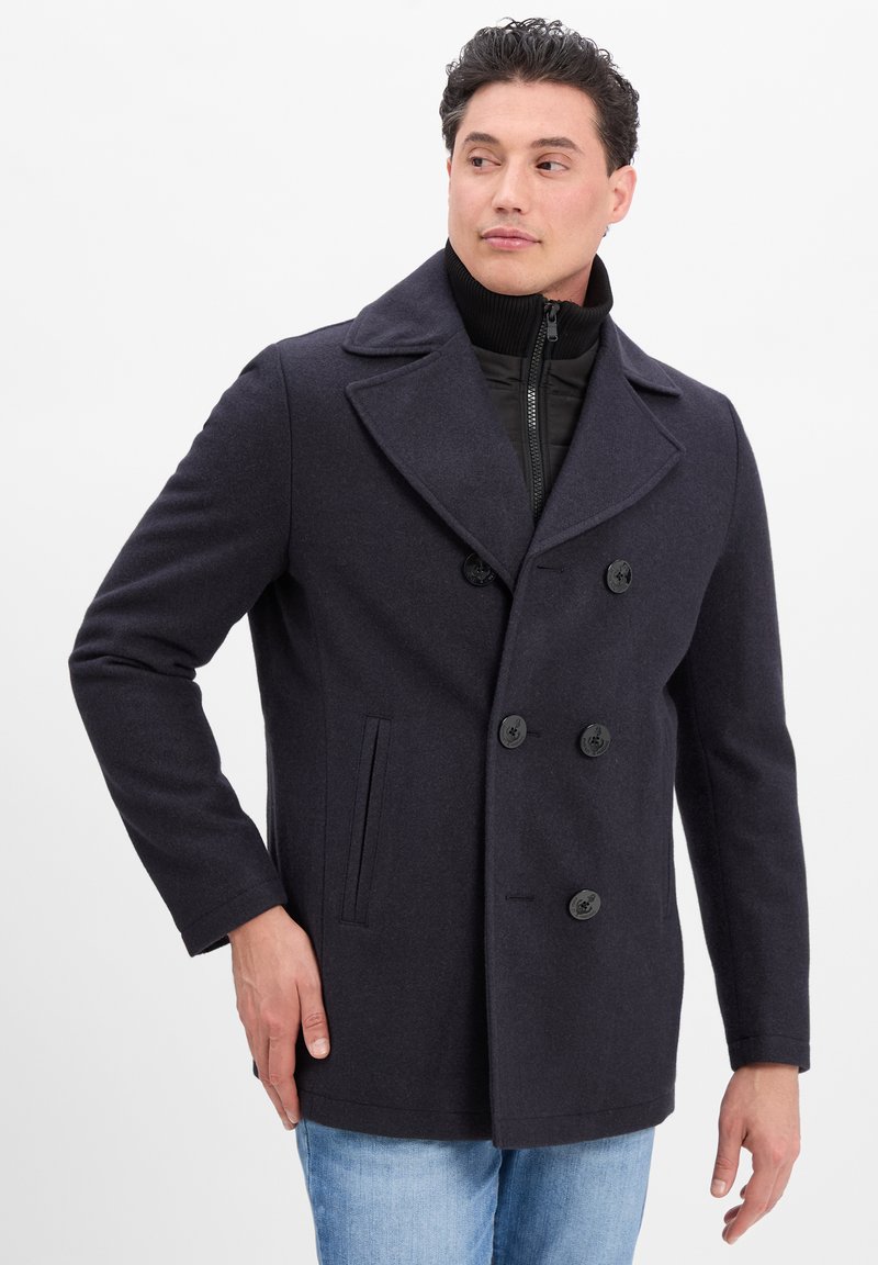 Mann trägt einen dunkelblauen, doppelt geknöpften Wollmantel über einem schwarzen Reißverschluss-Pullover und hellblauen Jeans und schaut zur Seite.