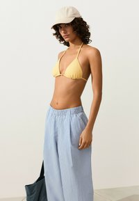 Jeune femme portant une casquette beige, un haut de bikini jaune et un pantalon ample rayé bleu, tenant un sac noir sur fond uni.