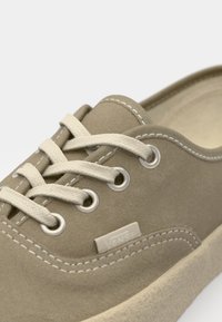 Beige zamšādas kurpes ar baltām auklām, metāla acīm un izšūtu apdari. Malā ir neliels Vans logotips. Apaļas purngalu dizains.