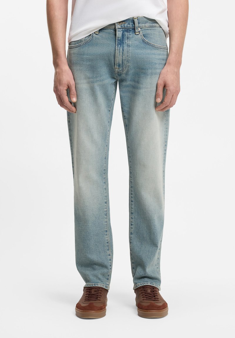 Άνδρας που φορά φωτεινό γαλάζιο ξεθωριασμένο τζιν και καφέ αθλητικά, στέκεται με τα χέρια χαλαρά στα πλάγια, μπροστά από ένα λευκό φόντο.