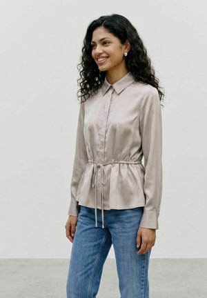 Femme souriante aux cheveux bouclés porte un chemisier beige à manches longues avec ceinture à la taille et un jean bleu, debout devant un fond clair uni.