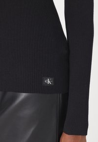 Närbild av en person som bär en svart ribbad tröja med en Calvin Klein-logotyp på en svart lädeskjol.