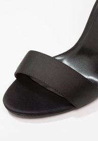 Sandale noire à bout ouvert avec une large sangle en tissu à l'avant, dotée d'une semelle intérieure lisse et brillante et d'un design minimaliste.