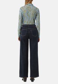 Hoge taille donkerblauwe denim wide-leg jeans met dubbele achterzakken en contrasterende stiksels, gecombineerd met een gemusterd langmouwtop in blauw en groen.