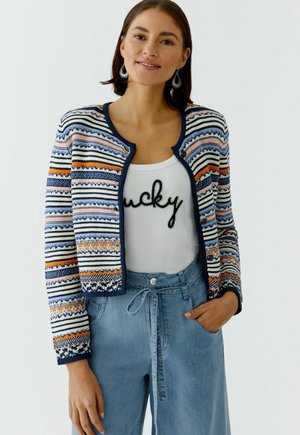 Vrouw draagt een meerkleurige gestreepte gebreide cardigan over een witte top met de tekst "lucky", gecombineerd met lichtblauwe jeans met hoge taille en trekkoord.