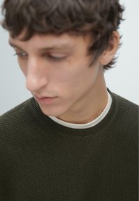 Maglione verde scuro con texture e collo rotondo, indossato sopra una maglietta bianca con scollatura tonda, mostrato in un primo piano del collo e dei dettagli del tessuto.