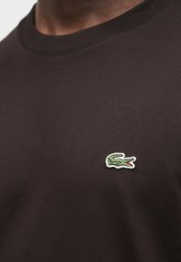 Άνδρας που φοράει μπλούζα καφέ σκούρου χρώματος με στρογγυλή λαιμόκοψη και μικρό κεντητό πράσινο λογότυπο κροκοδείλου στο αριστερό στήθος.