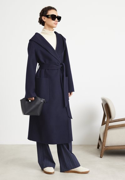 Cappotto avvolgente in lana blu navy con cappuccio, cintura in vita e grandi tasche, abbinato a pantaloni a righe sottili e sneakers bianche, mentre tiene una borsa nera.