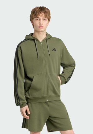 Ung mand iført en olivengrøn Adidas hættetrøje med lynlås og matchende shorts med sorte striber og logoer, stående med den ene hånd i lommen.