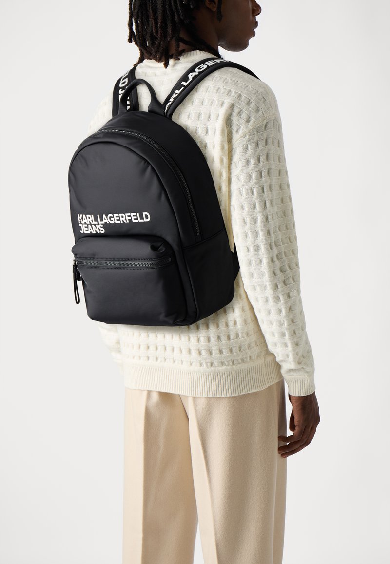 Mochila negra de nailon con forma redondeada, bolsillo frontal con cremallera y texto blanco "KARL LAGERFELD JEANS." Cuenta con correas dobles para los hombros.