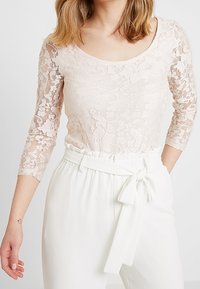 Spetsblus i ljusrosa med trekvartslånga ärmar, med ett blommönster och rundad halsringning, tillsammans med högmidjade vita byxor.