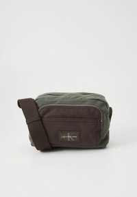 Axelremsväska i olivgrönt och brunt canvas med dragkedja, justerbar rem, ficka framtill och logotypmärke.