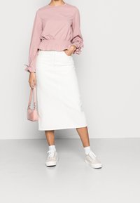 Camicia rosa chiaro a maniche lunghe con polsini a balze, abbinata a una gonna midi in denim bianco e sneakers beige. Include una piccola borsa rosa.