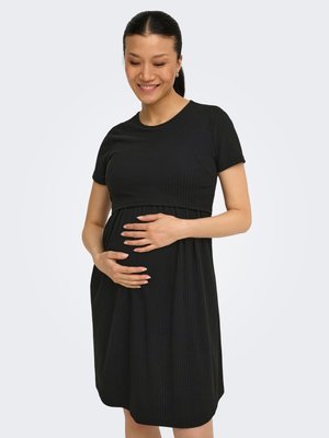 Femeie însărcinată în rochie neagră cu mâneci scurte care își ține ușor burta, zâmbind, stând în picioare pe un fundal deschis și simplu.