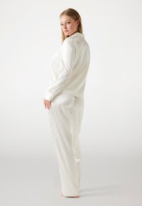 Weißes Satin-Pyjama-Set mit einem knöpfbaren Hemd und lockeren Hosen mit einem subtilen Blumenmuster. Glatte Textur mit einem sanften Glanz.