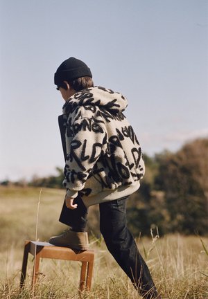 Child wearing a black beanie and patterned coat steps onto a wooden stool in a grassy outdoor area with clear sky.