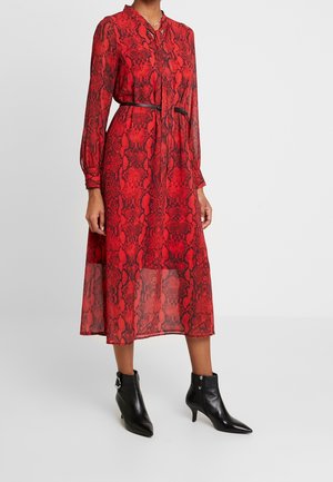 Vrouw die een midi-jurk met rood slangenprint draagt, met lange mouwen, een zwarte tailleriem en zwarte enkellaarsjes met een kleine hak en puntige neus.