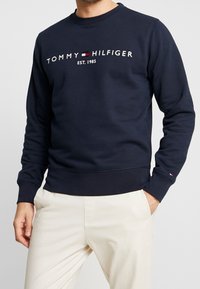 Hombre vestido con sudadera de Tommy Hilfiger azul marino, con texto del logo en blanco y pantalones color crema, con las manos en los bolsillos y el torso visible.