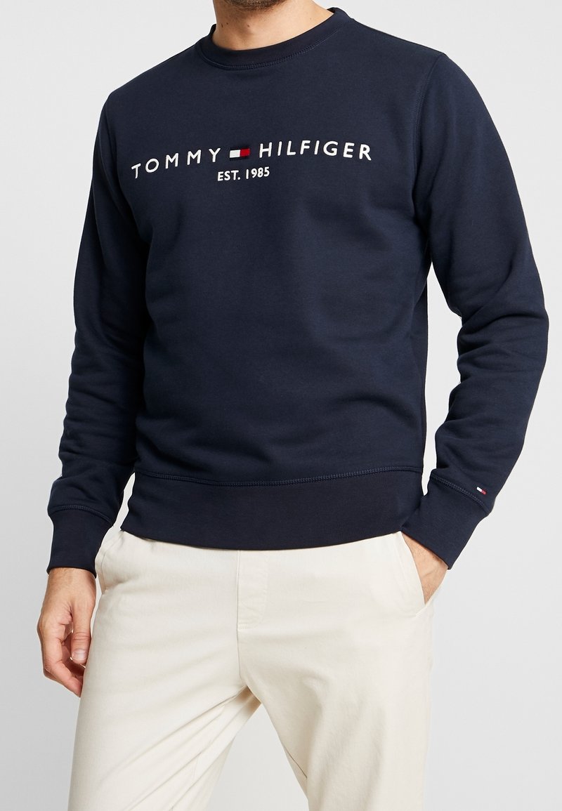 Uomo che indossa un felpa navy blue di Tommy Hilfiger con testo del logo bianco e pantaloni color crema, mani in tasca, torso visibile.