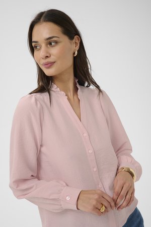 Frau mit langen dunklen Haaren, die eine hellrosa, geknöpfte Bluse mit Rüschkragen, goldene Creolen und goldene Ringe trägt und seitlich schaut.