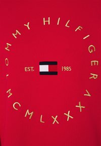 Rode stof met gouden borduursels "Tommy Hilfiger MCMLXXXV" rond een klein rechthoekig logo in rood, wit en zwart met "EST. 1985" in goud.