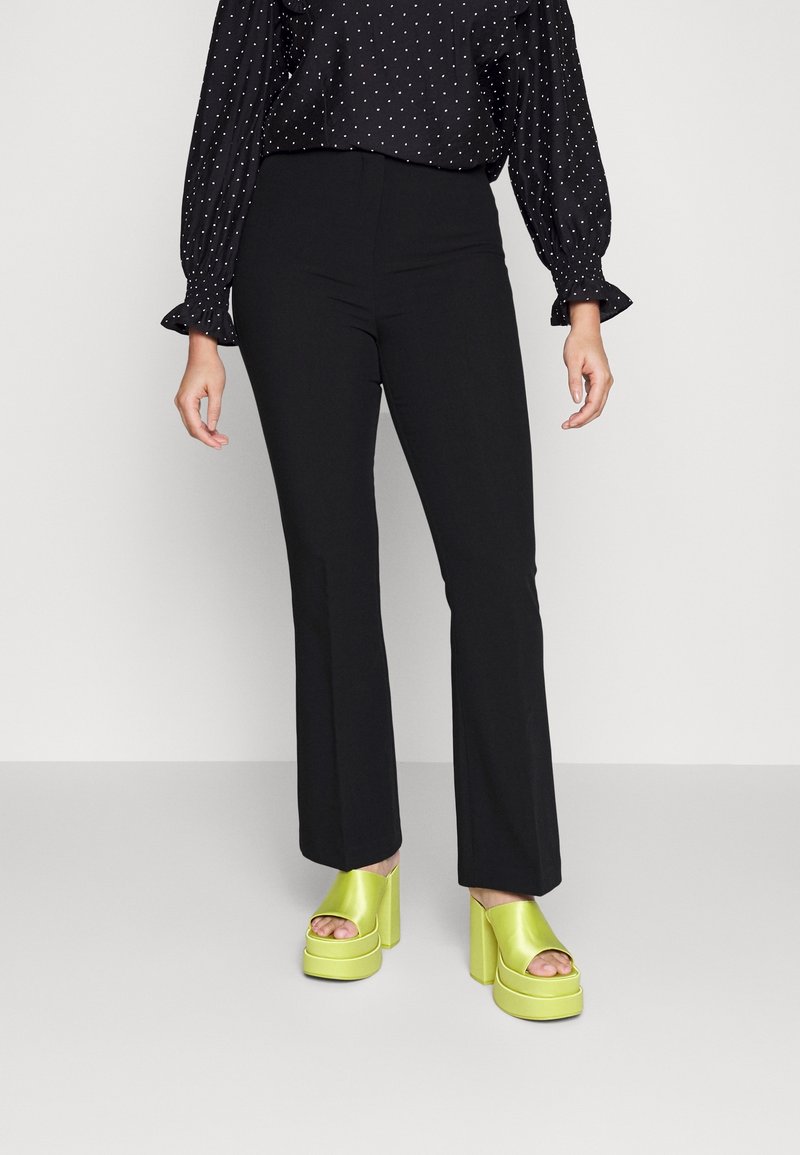 Zwarte hoogsluitende flare-broek gecombineerd met levendige limoengroene chunky platformhakken met een open teen ontwerp. Zwarte blouse met stippen erboven.