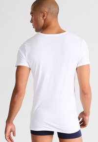 Muž v jednobarevném bílém tričku s krátkým rukávem a námořnickomodrých šortkách, viděný zezadu na jednobarevném světlém pozadí.