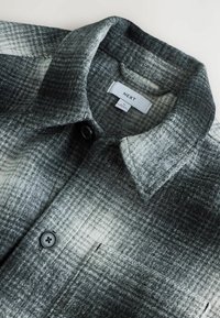 Camicia di flanella a quadri grigia e nera, con colletto a punta, chiusura con bottoni e tessuto texturizzato con un motivo delicato. Etichetta del brand visibile.