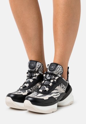 Jambes portant des baskets montantes Michael Kors à motif noir et blanc avec des semelles blanches épaisses sur un fond uni.