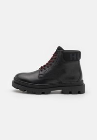Schwarze Leder-Ankle-Boots mit einer strukturierten Oberfläche, roten Schnürsenkeln und einem gepolsterten schwarzen Kragen mit "HU" in Blau. Dicke Gummisohle mit Profil.