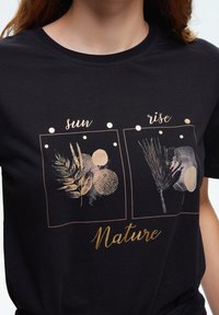 Czarna koszulka z nadrukami w kolorze złotym przedstawiającymi liście i koła, podpisanymi jako "słońce" i "wschód", z napisem "Natura" na dole.