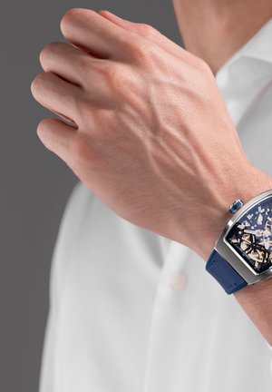 Moški zapestje z modrim pasom skeletne ure, roka dvignjena blizu obraza, oblečen v belo srajco na sivem ozadju.